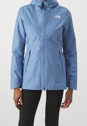 Vrouw draagt een lichtblauwe jas met capuchon, een rits aan de voorkant en met ritsen aan de zijzakken, gecombineerd met zwarte leggings.