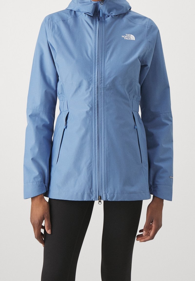 Femme portant une veste à capuche bleu clair avec une fermeture éclair à l’avant et des poches latérales zippées, associée à des leggings noirs.