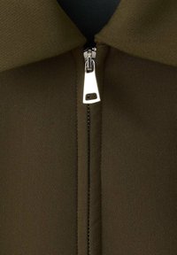 Oliivinvihreä kangas, jossa on sileä pinta, keskellä hopeinen vetoketju ja vetoketjun yläpuolella kaulussuunnittelu.