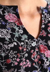 Sorte stof med blomstermønster i forskellige farver, herunder rød, pink, blå og grøn; har en V-hals-design og let tekstur.
