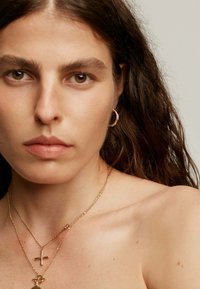 Pendiente de aro de oro, collares de oro en capas con diferentes texturas de cadena y un colgante de cruz, sobre un fondo neutro.