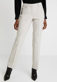 Vaaleanbeige muotihousut sileällä pinnalla, joissa on litteät etutaskut ja kapea suora lahje, yhdistetty mustiin korkokenkiin.