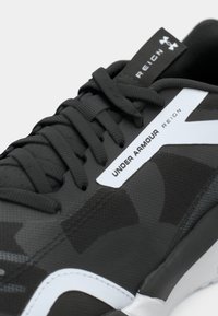 Μαύρο και γκρι αθλητικό παπούτσι με αναπνεύσιμο επάνω μέρος, υφασματικά μοτίβα, λευκές λεπτομέρειες και κορδόνια. Φέρει το λογότυπο «Under Armour».