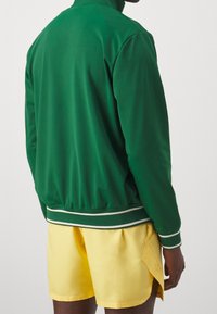 Groene ritsjack in een gladde stof met witte accenten, gecombineerd met gele shorts, die een ontspannen pasvorm en casual ontwerp tonen.