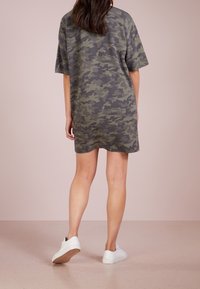 Szara sukienka w kamuflażu w kształcie T-shirtu z krótkimi rękawami, luźny krój i zaokrąglone wykończenie. W połączeniu z białymi sneakersami na gładkiej, jasnej podłodze.
