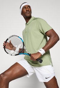 Grønn polo med korte ermer og en logo, kombinert med hvite shorts. Spilleren holder en tenniskølle med blå detaljer, iført et hvitt hodebånd.