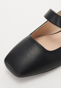 Chaussure Mary Jane en cuir noir avec un bout carré, une sangle sur le cou-de-pied et une texture lisse. L'intérieur présente un revêtement beige.