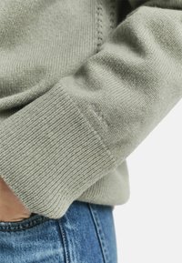 Maglione verde chiaro in maglia con polsini a coste e un logo discreto. La texture del tessuto appare morbida e calda, abbinata a denim blu.