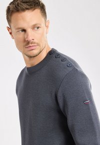 Marineblauer Strickpullover mit Rundhalsausschnitt, der dekorative Knöpfe an der Schulter und ein kleines Logo am Ärmel aufweist. Sanfte Textur.