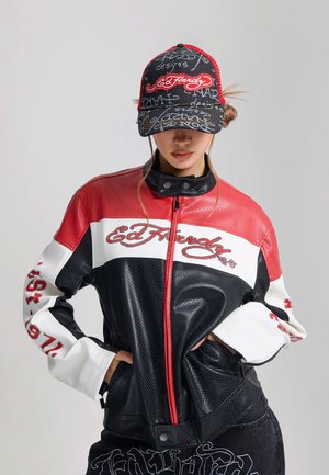Persona vistiendo una chaqueta Ed Hardy negra, roja y blanca y gorra a juego con texto de la marca y detalles gráficos, manos en los bolsillos de la chaqueta.
