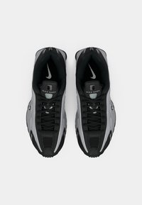 Crni i sivi Nike Shox tenisice s mrežastim gornjim dijelom, teksturiranim potplatom i istaknutim sustavom vezivanja uz reflektirajuće naglaske.