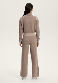 Tuta in maglia marrone chiaro con colletto alto, polsini a coste e pantaloni a gamba larga con spacco laterale. Tessuto morbido e design casual.