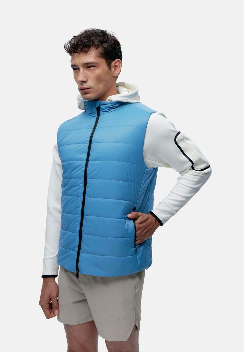 Gilet isolant bleu clair avec motif matelassé, fermeture éclair noire, deux poches latérales, porté par-dessus une chemise à manches longues blanche avec des accents noirs.