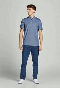 Camisola polo azul com uma pala de três botões e mangas curtas, combinada com jeans escuros e ténis brancos, de pé contra um fundo claro.