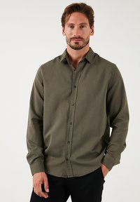 Camisa verde oliva de botones, hecha de un tejido texturizado, con cuello, mangas largas y botones negros. Llevada por un modelo frente a un fondo neutro.