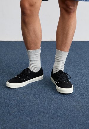 Black suede sneakers with white rubber soles, adorned with decorative metal studs. Worn with light gray ribbed socks on a blue carpet.