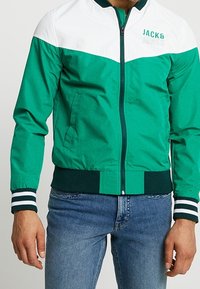 Grön och vit zipjacka med elastiska muddarna och nederkant. Har "JACK & JONES"-logotypen på vänster sida. Tillverkad av ett glatt, lätt material.