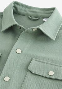 Camisa de botones verde claro con un acabado texturizado, que cuenta con dos bolsillos frontales y detalles en hardware plateado en los botones.