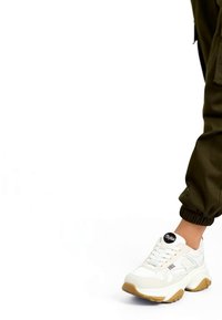 Rechterbeen met olijfgroene joggerbroek en witte chunky sneaker met gumzool op witte achtergrond.