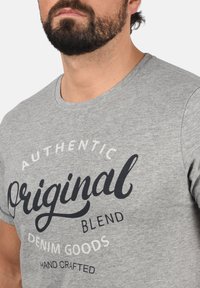 Pelēkā kokvilnas T-krekls ar apaļu kakla izgriezumu un melnbaltu ekrāndrukātu tekstu, kas vēsta "Autentiskas oriģinālas samaisa džinsas preces, roku darbs".