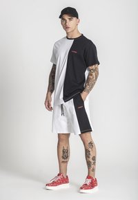 Tričká s krátkym rukávom v čierno-bielom farebnom bloku, spárované s bielymi šortkami s čiernymi akcentmi a červeným nápisom; červené tenisky s vzorom.