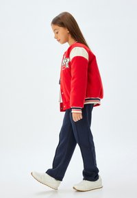 Rød og hvid varsityjakke med marineblå træningsbukser og hvide sneakers. Jakken har logo, knapper og stribede manchetter. Glat stof.