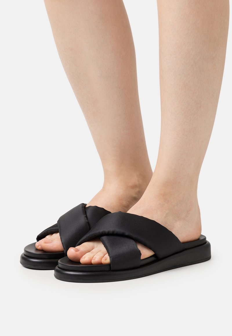 Pieds portant des sandales slip-on noires rembourrées à brides croisées avec semelles plates sur fond blanc uni.