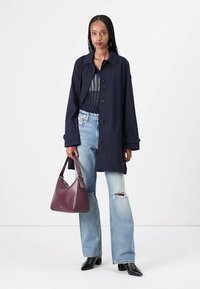Marine trenchcoat med store knapper, kombinert med lyseblå revnejeans. Holder en bordeaux lærveske med glatt tekstur. Svarte spisse sko.