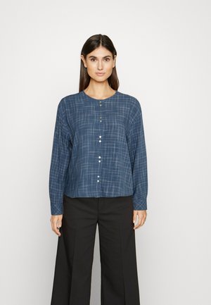 Ναυτικό μπλε πουκάμισο με κουμπιά, με καρό σχέδιο, μακριά μανίκια, στρογγυλή λαιμόκοψη και δύο πράσινα κουμπιά στο λαιμό. Συνδυάζεται με μαύρες παντελόνες φαρδιές.