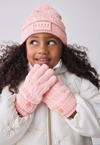 Lyserøde strikkede handsker har et kabelmønster med perle-accents og knappedetaljer. En matchende lyserød strikhue med en pelskvast bæres.