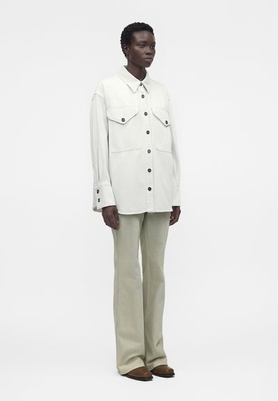 Άτομο που φοράει υπερμεγέθη άσπρο πουκάμισο με τσέπες με καπάκια και ανοιχτό μπεζ φαρδιά παντελόνια, στέκεται μπροστά σε απλό φόντο.