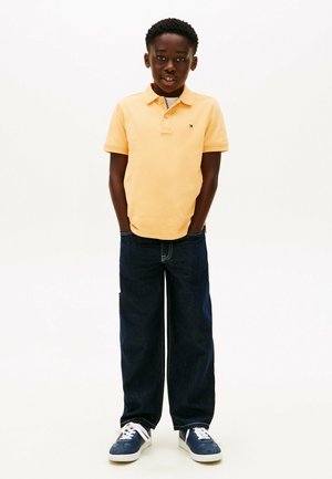 Ung dreng stående med hænderne i lommerne iført en lyseorange poloshirt, mørkeblå jeans og blå sneakers på en hvid baggrund.