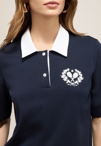 Námořnické polo tričko s bílým límečkem, zapínáním na tři knoflíky a vyšitým tenisovým motivem obklopeným vavřínovými listy na hrudi.