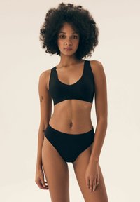 Kobieta z kręconymi włosami, ubrana w czarny stanik i majtki z wysokim stanem, stojąca na tle jasnej, jednolitej ściany z widocznymi tatuażami na ramionach.