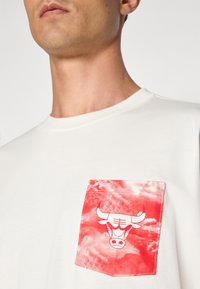Hvid T-shirt med en rød lomme og et kvæg grafik på en skyet baggrund. Glat stof med rund halsudskæring.