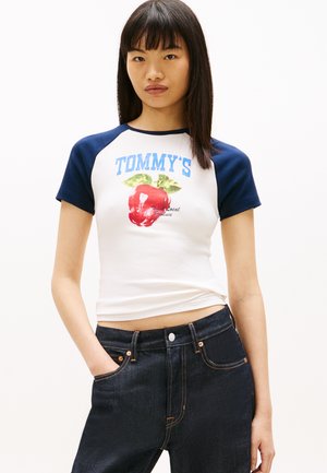 Giovane donna con capelli castani lisci indossa una maglietta a maniche corte bianca e blu navy con la scritta "TOMMY'S" e un'illustrazione di una mela rossa, abbinata a jeans scuri.