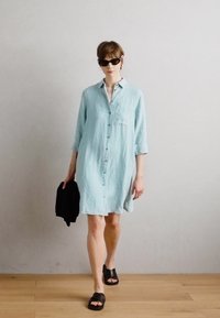 Ανοιχτό μπλε, πουκάμισο φόρεμα από λινό με γυρισμένα μανίκια και μπροστινή τσέπη. Συνδυάζεται με μαύρες παντόφλες. Κρατάει μια μαύρη ζακέτα.