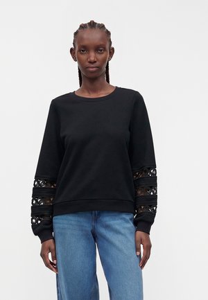 Kvinde iført en sort langærmet top med blondeudskæringer på ærmerne og blå jeans, stående mod en ensfarvet lys baggrund.