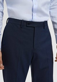 Pantalon bleu marine au design épuré, avec une fermeture à bouton, des passants de ceinture et un tissu texturé, présenté avec une chemise bleu clair.