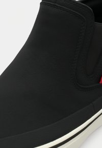 Black slip-on shoe featuring a smooth texture, elastic side panels, and a white rubber sole. Notable red accent on the inner lining.