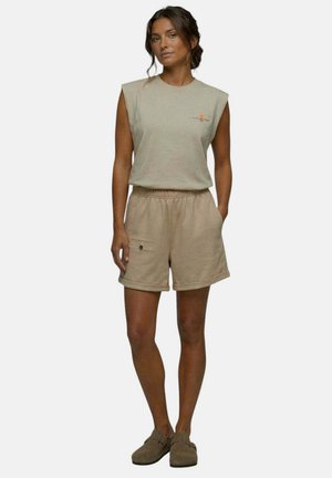 Femme aux cheveux bruns en chignon bas porte un haut beige sans manches, un short assorti avec taille élastique et poche, ainsi que des chaussures marron à enfiler, debout, détendue.