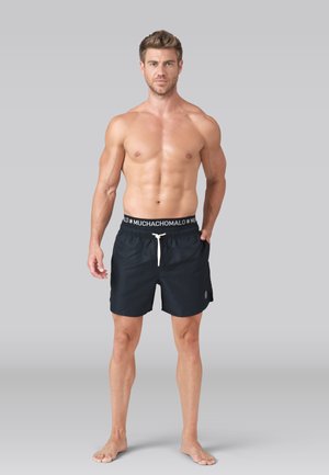 Homme en forme debout pieds nus portant un short de bain noir avec cordon blanc et taille élastique de marque sur fond gris.