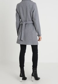 Cappotto di lana grigio con cintura, vestibilità aderente, tasche a soffietto e maniche lunghe. Abbinato a jeans neri e stivaletti con tacco argentato.