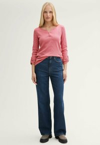 Chemise henley rose à manches longues avec boutons, associée à un jean évasé bleu foncé et des chaussures gris à enfiler, sur un fond neutre.