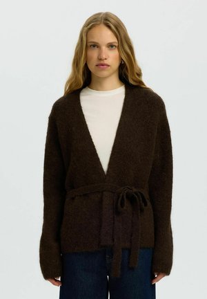 Jeune femme aux longs cheveux blonds porte un cardigan marron foncé et duveteux noué à la taille sur un haut blanc et un jean bleu.