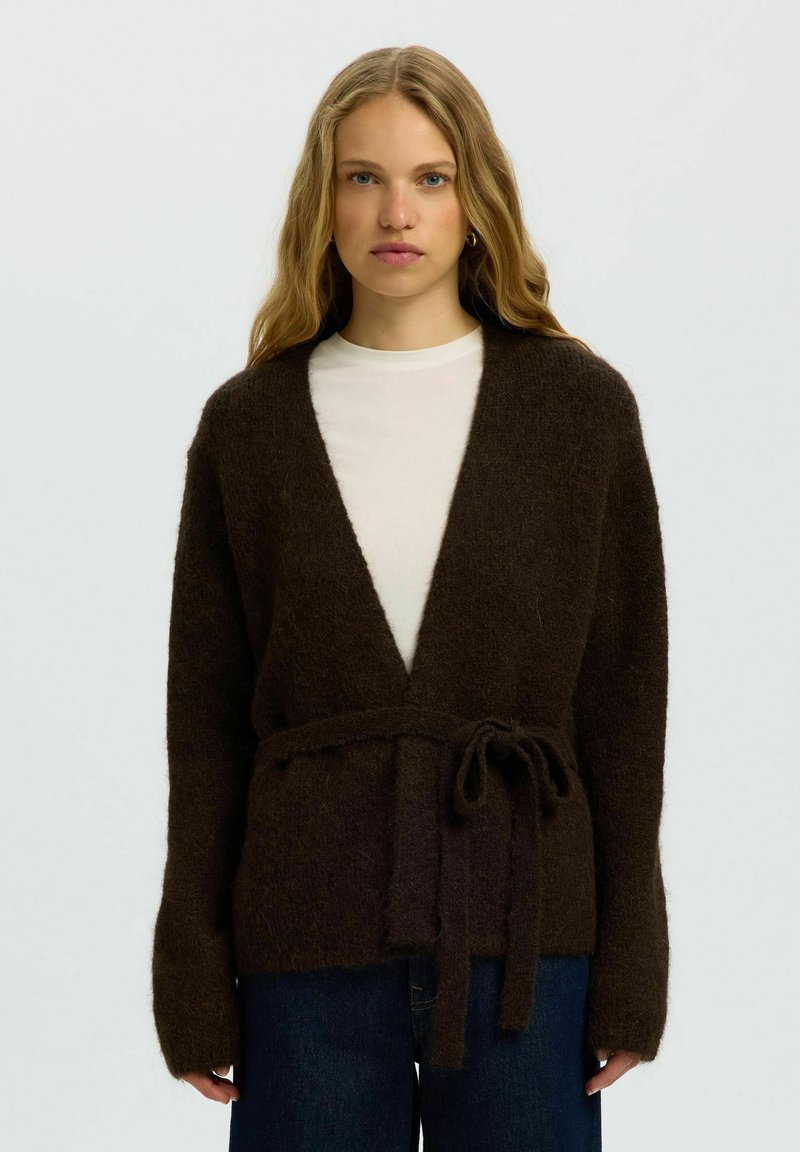 Jeune femme aux longs cheveux blonds porte un cardigan marron foncé et duveteux noué à la taille sur un haut blanc et un jean bleu.