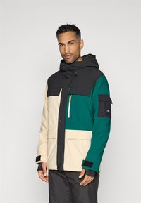 Casaco impermeável em blocos de cores, apresentando painéis nas cores preto, verde e creme. Inclui capuz, bolsos com fecho éclair e fechos de Velcro nos punhos.