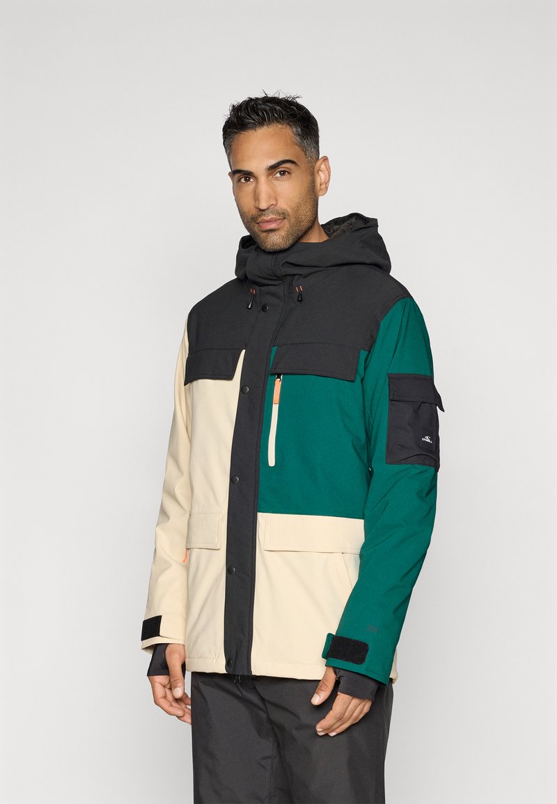 Spalvotai blokų iš vandeniui atsparaus audinio pagaminta striukė, turinti juodus, žalius ir kreminius segmentus. Su gobtuvu, užtraukiamomis kišenėmis ir Velcro užsegimais ant rankogalių.