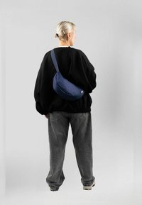 Borsa a tracolla blu con forma arrotondata, materiale in tela, ampia tracolla e piccolo etichetta del marchio. Indossata sopra un maglione nero oversize e jeans grigi.