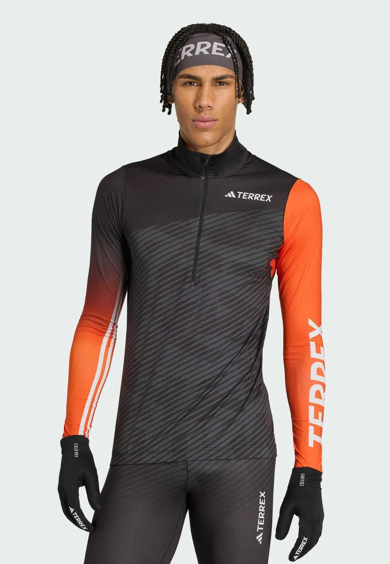 Athlète masculin portant un haut Terrex à manches longues noir et orange, des gants noirs et un bandeau, debout devant un fond gris clair.